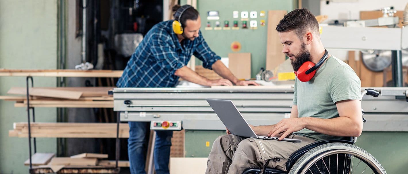 Ein Mann im Rollstuhl arbeitet in einer Schreinerei an einem Laptop, den er auf dem Schoß hat. Im Hintergrund ist sein Kollege zu sehen.